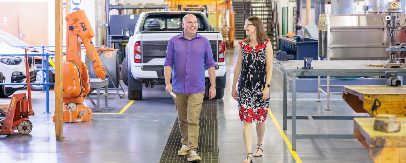 Two people walking across a workshop floor past a robot arm and a pickup truck