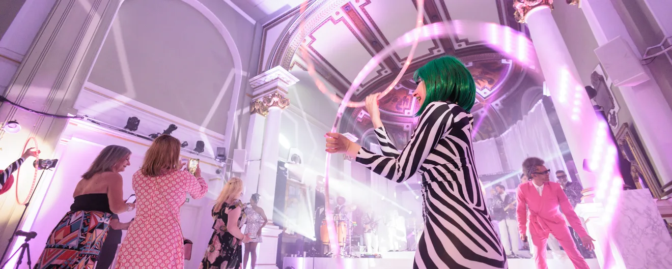 A performer in a striped catsuit spinning a light hoop among guests under a pink-lit ceiling