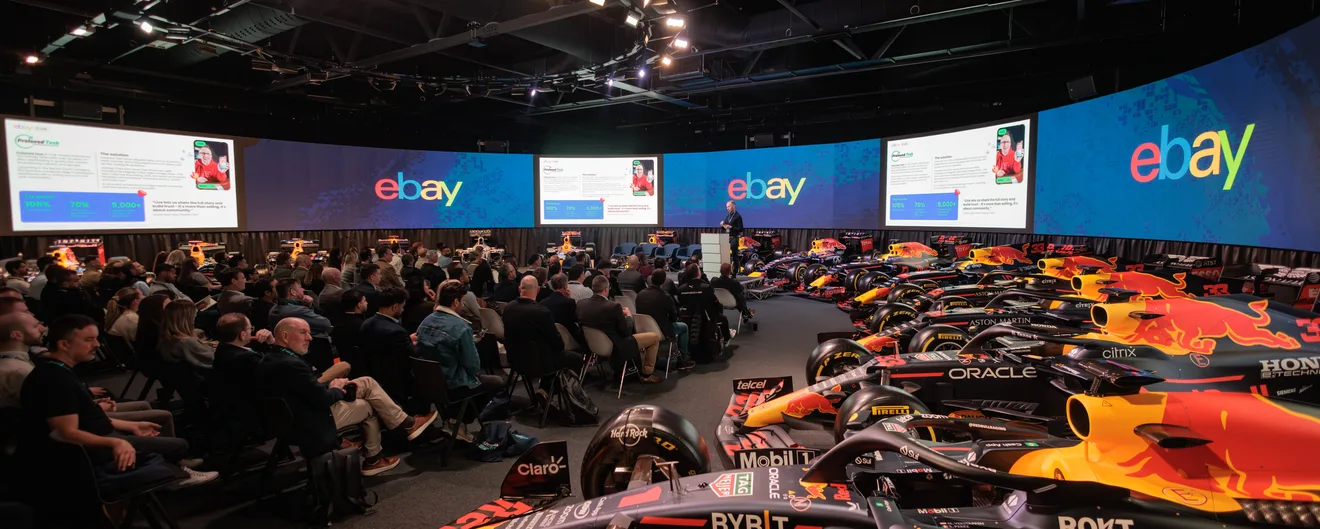 A conference audience beneath a curved wall of screens, with racing cars on display