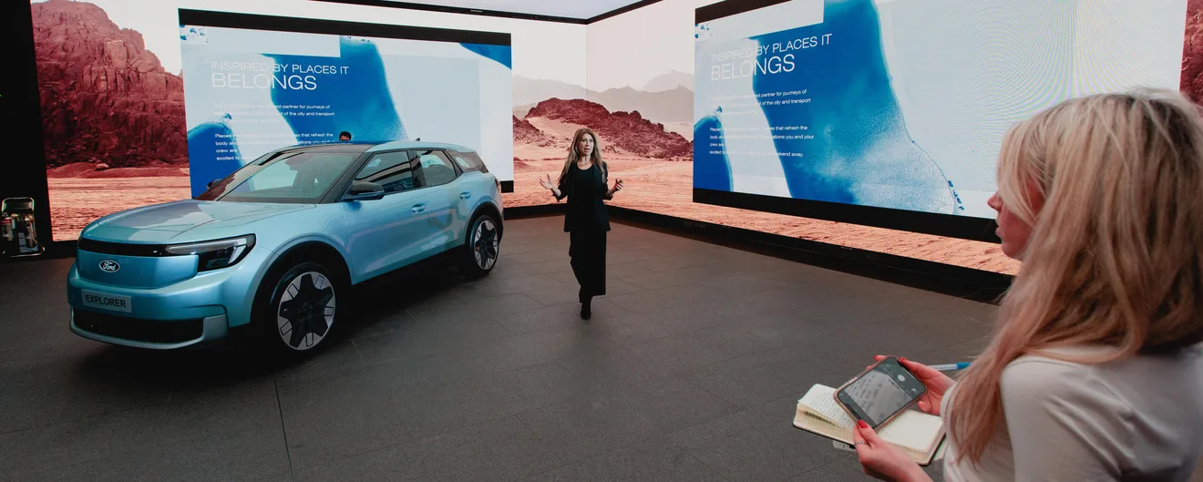 A presenter walking past a new Ford car in front of a wide screen of desert landscapes
