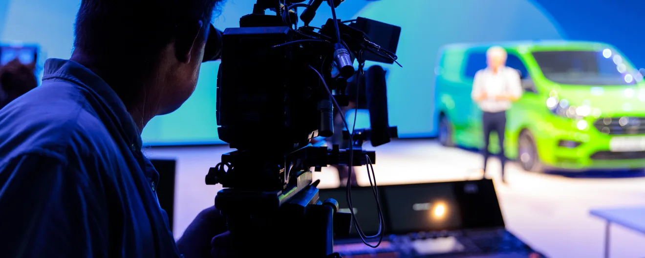 A camera operator filming a presenter beside a green Ford E-Transit on stage
