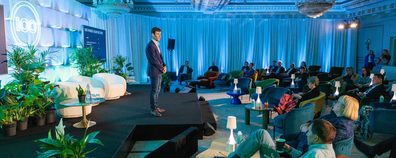A speaker on stage before a seated audience in a blue-lit ballroom